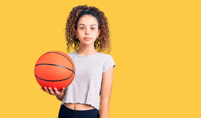 Beautiful kid girl with curly hair holding basketball ball thinking attitude and sober expression looking self confident