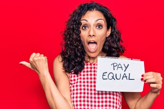 Middle age beautiful woman holding pay equal paper pointing thumb up to the side smiling happy with open mouth