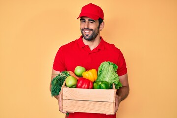 Handsome hispanic man with beard wearing uniform holding delivery food winking looking at the...