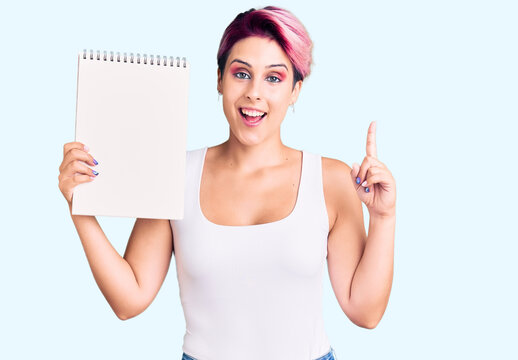 Young beautiful woman with pink hair holding notebook surprised with an idea or question pointing finger with happy face, number one