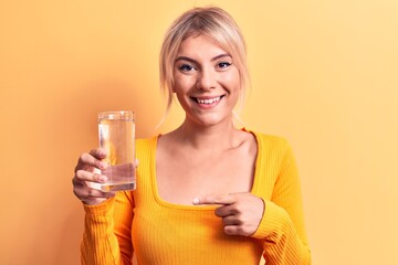 Young beautiful blonde woman drinking glass of water over isolated yellow background smiling happy pointing with hand and finger