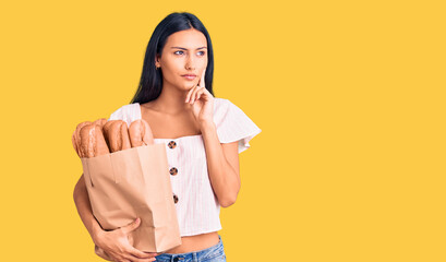 Young beautiful latin girl holding paper bag with bread serious face thinking about question with hand on chin, thoughtful about confusing idea