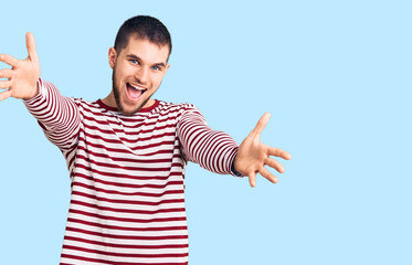 Young handsome man wearing striped sweater looking at the camera smiling with open arms for hug. cheerful expression embracing happiness.