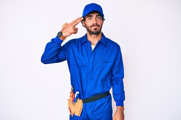 Handsome young man with curly hair and bear weaing handyman uniform shooting and killing oneself pointing hand and fingers to head like gun, suicide gesture.