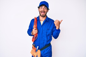 Handsome young man with curly hair and bear wearing builder jumpsuit uniform and electric cables pointing thumb up to the side smiling happy with open mouth