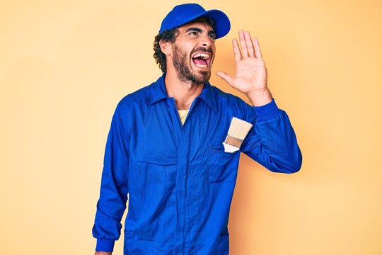 Handsome young man with curly hair and bear wearing builder jumpsuit uniform shouting and screaming loud to side with hand on mouth. communication concept.