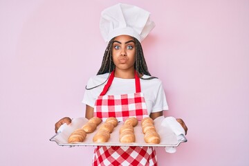 Young african american woman with braids wearing baker uniform holding homemade bread puffing cheeks with funny face. mouth inflated with air, catching air.