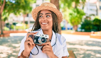 Young african american woman with braids smiling happy taking pictures with vintage camera outdoors on a sunny day of summer