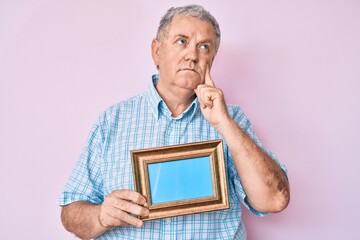 Senior grey-haired man holding empty frame serious face thinking about question with hand on chin,...