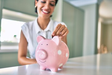 Young brunette woman smiling happy putting money savings inside of piggy bank