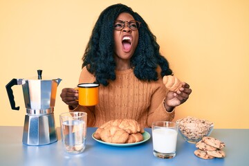 Young african american woman eating breakfast holding croissant angry and mad screaming frustrated and furious, shouting with anger. rage and aggressive concept.