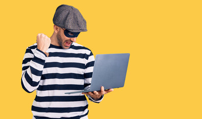 Young handsome man wearing burglar mask using laptop screaming proud, celebrating victory and...