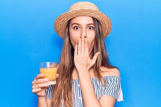 Young beautiful girl wearing summer hat drinking glass of orange juice covering mouth with hand, shocked and afraid for mistake. surprised expression