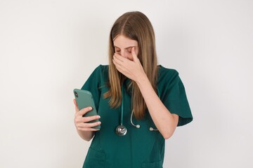A  Female doctor wearing a green scrubs and stethoscope looking at smart phone feeling sad holding...