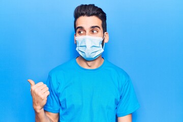 Young handsome man with beard wearing medical mask pointing thumb up to the side smiling happy with open mouth
