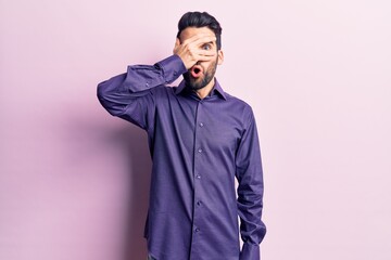 Fototapeta premium Young handsome man with beard wearing casual shirt peeking in shock covering face and eyes with hand, looking through fingers with embarrassed expression.