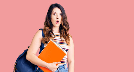 Beautiful young brunette woman wearing student backpack hodling books scared and amazed with open...