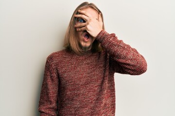 Handsome caucasian man with long hair wearing casual winter sweater peeking in shock covering face and eyes with hand, looking through fingers afraid