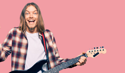 Handsome caucasian man with long hair playing electric guitar screaming proud, celebrating victory and success very excited with raised arms