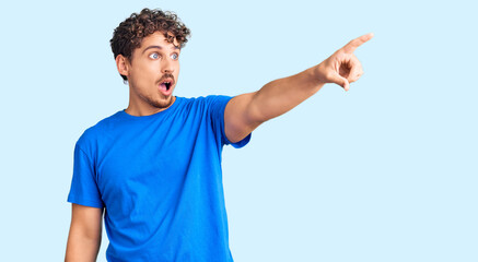 Young handsome man with curly hair wearing casual clothes pointing with finger surprised ahead, open mouth amazed expression, something on the front