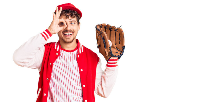 Young handsome man with curly hair wearing baseball uniform holding golve and ball smiling happy doing ok sign with hand on eye looking through fingers