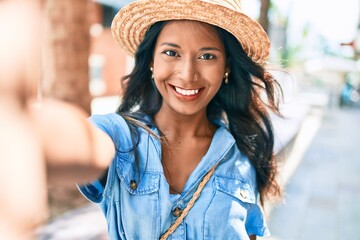 Young beautiful indian woman wearing summer hat smiling happy making selfie by the camera at the city.