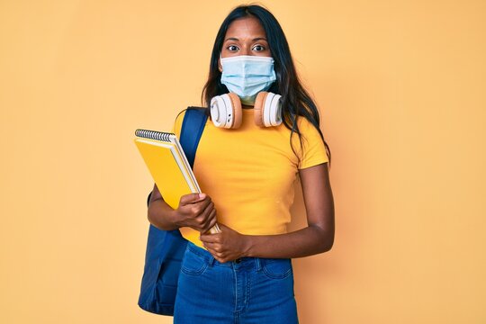 Young Indian Girl Wearing Medical Mask Holding Student Backpack And Books Scared And Amazed With Open Mouth For Surprise, Disbelief Face