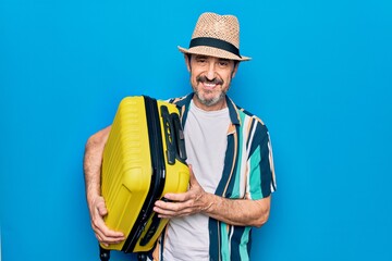 Middle age handsome tourist man on vacation holding cabin bag over isolated blue background looking positive and happy standing and smiling with a confident smile showing teeth