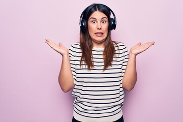 Young beautiful brunette woman listening to music using headphones over pink background clueless and confused with open arms, no idea and doubtful face.
