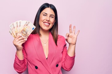 Young beautiful businesswoman holding euros banknotes over isolated pink background doing ok sign with fingers, smiling friendly gesturing excellent symbol