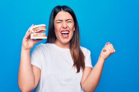 Young beautiful brunette woman asking for dental care holding denture with backets screaming proud, celebrating victory and success very excited with raised arm