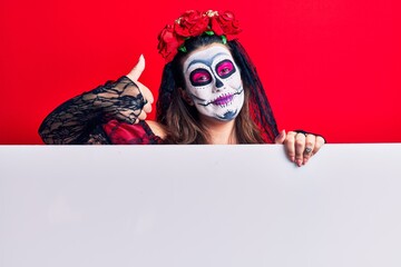 Young woman wearing day of the dead costume holding blank empty banner smiling happy and positive, thumb up doing excellent and approval sign