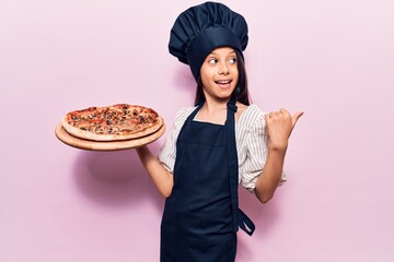 Beautiful child girl wearing cooker uniform holding italian pizza pointing thumb up to the side smiling happy with open mouth