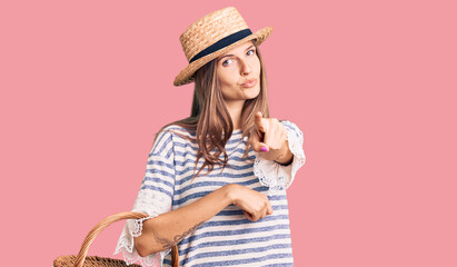 Beautiful caucasian woman wearing summer hat and holding picnic wicker basket with bread pointing...