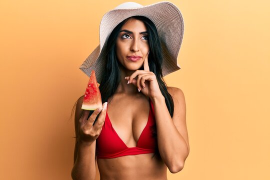 Beautiful hispanic woman wearing bikini eating watermelon serious face thinking about question with hand on chin, thoughtful about confusing idea
