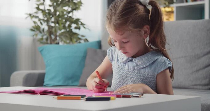 Happy little kid in blue dress drawing with colorful pencils in album. Smiling girl with two ponytails developing creativity during free time at home.