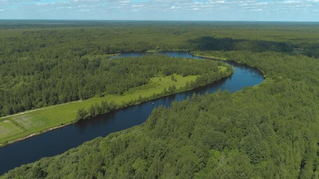 Aerial view of the bend of the river surrounded by green fields through which the river flows. Shot. Natural landscape of the river curve and lush green trees on blue cloudy sky background.