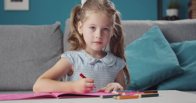 Pretty girl in blue dress using colorful pencils for painting in drawing-book on table, smiling and looking at camera. Concept of childhood and activity.