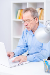 Elderly senior man with glasses on his face works remotely at home while sitting at a laptop, looking intently at the computer screen. Job for seniors during the coronavirus pandemic