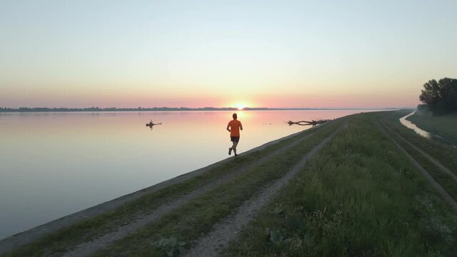 Young athlete, jogging alone on the lakeside track during golden hour. Follow through action shot from back.