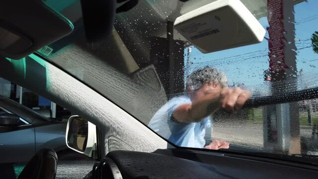 POV car passenger: Caucasian adult woman cleaning window windshield outside vehicle at gas station squinting on bright sunny blue sky day, handheld close up portrait