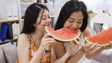 Best girl friends eating watermelon on sofa. Cheerful young women having fun biting fresh fruit tasty smiling laugh summer at home joyful moment day. Concept of healthy seasonal food shared together.