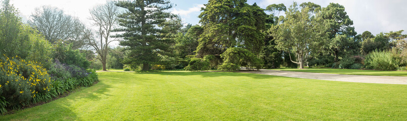 Panoramic view of a large formal garden landscaping with well-tended neat lawn and a variety of trees, flower beds and walkway. Background texture of grass and tall trees in a park on a sunny day.