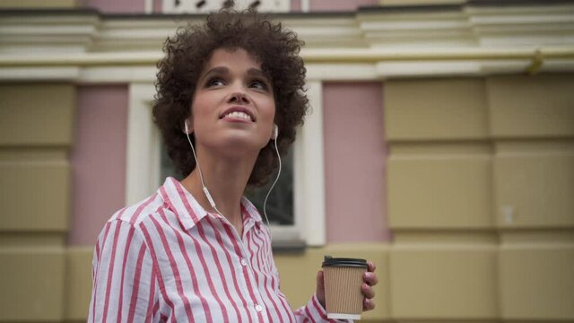 Happy smiling young adult hipster woman tourist wearing earphones drinking coffee listening music or audio travel tour guide in app enjoying sightseeing walking on urban city street outdoors.