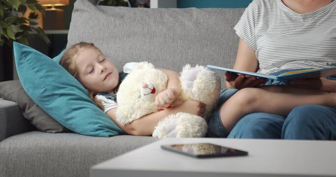 Lovely mother reading book to her little daughter before sleep. Pretty girl lying on grey couch hugging white teddy bear. Concept of careness.