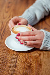 Young woman holding cup of coffee with latte art. She is wearing gray wool sweater and trendy silver ring. Morning in a cafe.