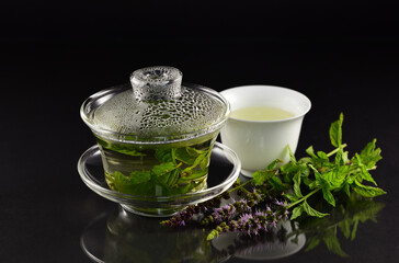 In a Chinese Gaiwan made of glass there is fresh warm peppermint tea in front of a dark background...