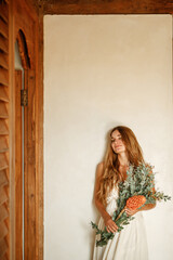 Young woman with eyes closed stand near the wall, holding a bouquet of flowers. Boho wedding style