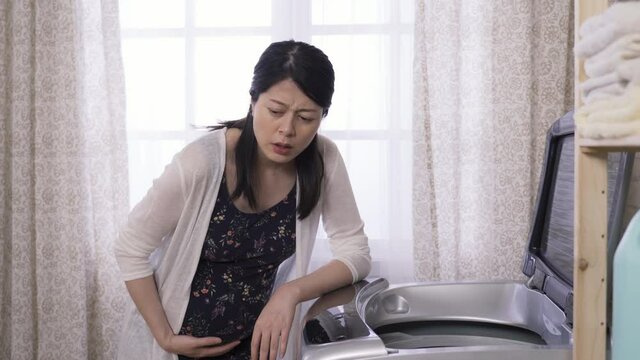 Asian housewife standing and resting by the washer at home is stroking abdomen, and wearing painful expression is having signs of labor.