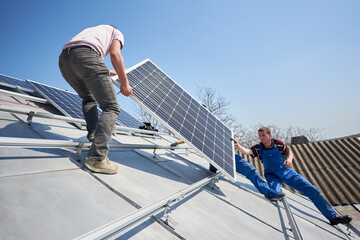 Male workers installing stand-alone solar photovoltaic panel system. Electricians lifting blue solar module on roof of modern house.